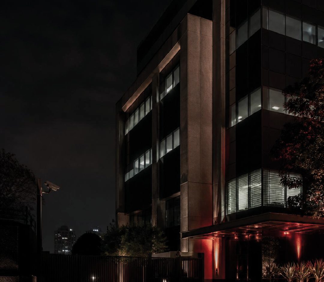 Fachada de edifício empresarial à noite, com portão fechado e iluminação em tons de vermelho.